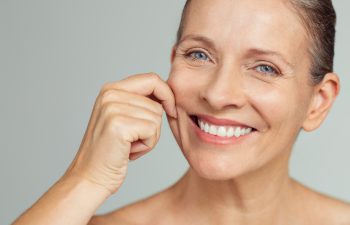 Senior woman pulling cheeks to feel softness and looking at camera. Beauty portrait of happy mature woman smiling with hands on cheek isolated over grey background. Aging process and skin concept.