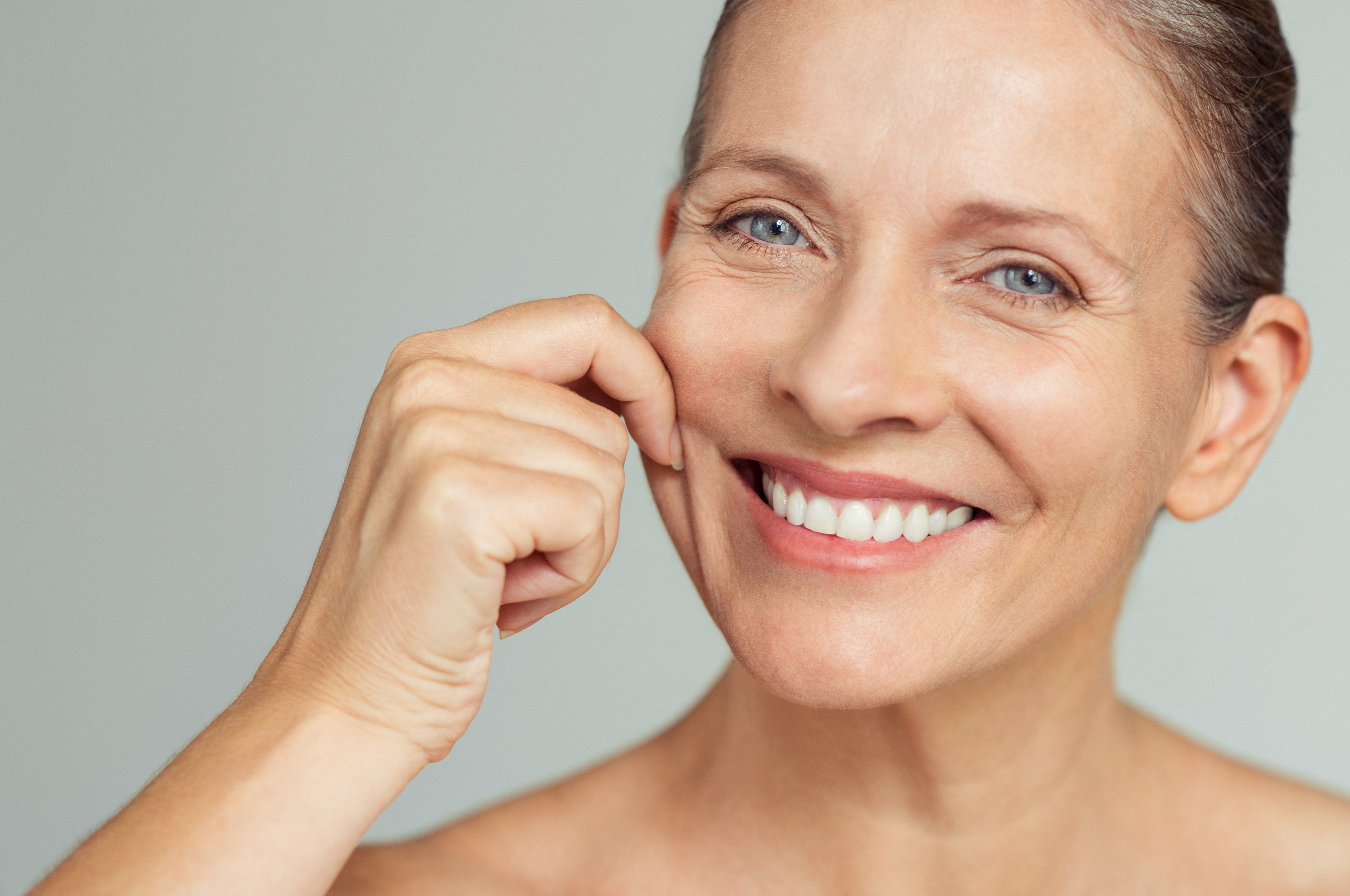 Senior woman pulling cheeks to feel softness and looking at camera. Beauty portrait of happy mature woman smiling with hands on cheek isolated over grey background. Aging process and skin concept.