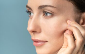 Young woman without makeup touching cheeks after glycolic acid peel, has signs of aging skin on her face, close up, isolated on studio blue background with copy space.